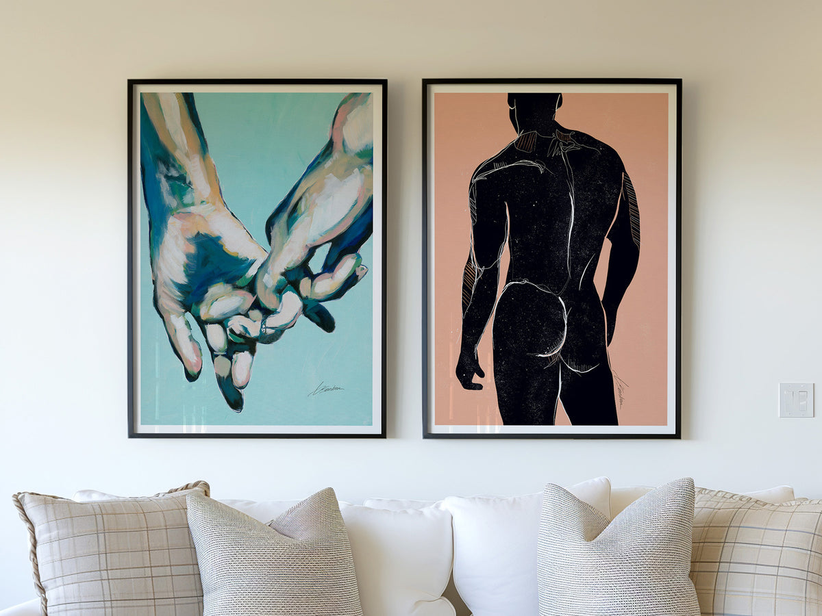 Two framed art prints by Brenden Sanborn above a white sofa: clasped hands on teal and a figure's back in black on peach