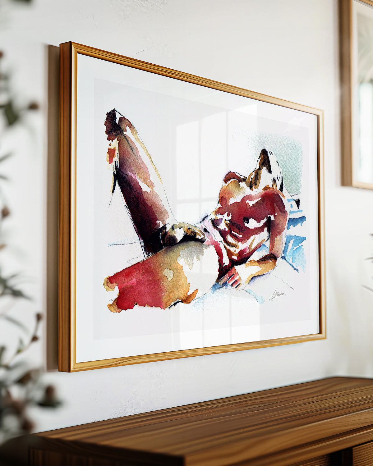 Lazy Light art print by Brenden Sanborn, a nude man lying back on blue sheets with one knee raised, in red and gold washes, shown in a light wood frame on a sideboard