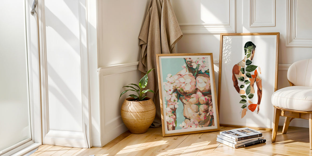 Two framed male figure art prints by Brenden Sanborn, a floral watercolor torso and a botanical leaf figure, leaning against a white paneled wall in a sunlit living room